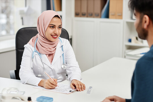 Medium shot of smiling Muslim female doctor consulting male patient in modern clinic office