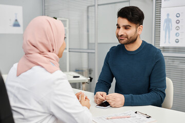 Medium shot of smiling male Muslim patient listening to female doctor wearing headscarf during consultation in clinic - obrazy, fototapety, plakaty