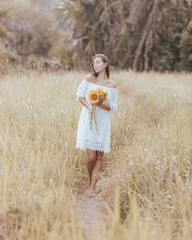 Romantic woman walking in a field and holding a bouquet of sunflowers. Charming Caucasian woman wearing white dress. Summer time. Lifestyle concept.