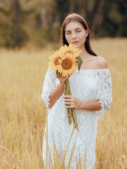 Caucasian woman walking in a field and holding a bouquet of sunflowers. Portrait of charming woman wearing white dress. Summer vacation. Lifestyle concept. Romantic mood.