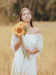 Caucasian woman walking in a field and holding a bouquet of sunflowers. Portrait of beautiful woman wearing white dress. Summer vacation. Lifestyle concept. Romantic mood.