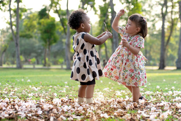 Two cute little girls with different skin colors playing at the park, pick up fallen flowers that...