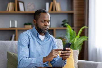 Serious thinking mature man sitting on sofa at home, senior african american man holding phone,...