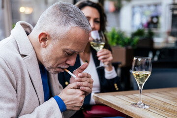 Senior man smoking a cigarette at a terrace while having a drink. Middle aged man smoking tobacco and drinking wine in a terrace with a woman.