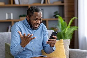 Sad nervous man sitting at home on sofa in living room of house, African American man received bad...