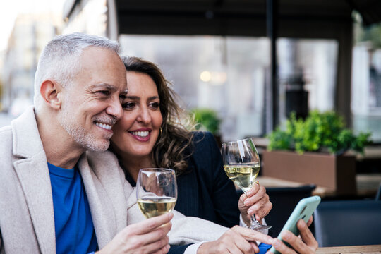 Happy senior couple sharing smartphone while drinking wine in city eating lunch at outdoors bar. Joyful mature man and woman using mobile phone together at a restaurant terrace.