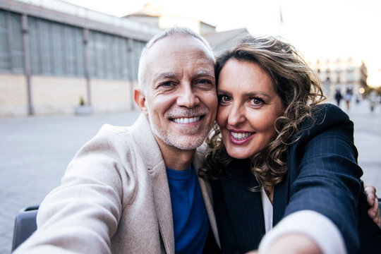 Affectionate mid adult happy couple having fun taking a selfie portrait together on the street. Middle aged man and woman friends smiling looking at camera.