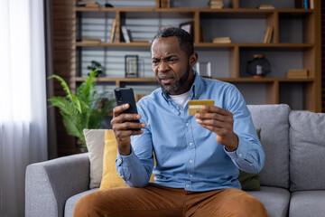 Upset and sad man sitting at home on couch in living room, cheated senior mature african american man displeased holding phone and bank credit card, rejected fund transfer error, account block. - obrazy, fototapety, plakaty