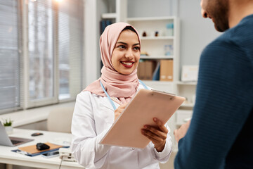 Portrait of smiling Muslim female physician in pink headscarf holding clipboard and welcoming unrecognizable male patient coming for consultation in medical center