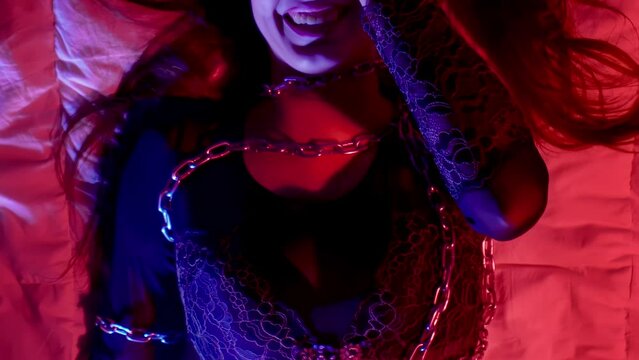 BDSM fetish concept, sexy shy bashful girl in erotic lingerie tied up with metal chains lying on bed, temptation. Hot passionate sensual woman. Intimate adult games with punishment. Brunette laughs