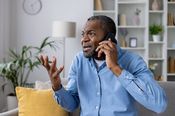 Angry nervous man sitting on sofa at home talking on the phone, unhappy african american man...