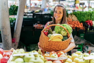 A portrait of a smiling Caucasian girl holding a basket filled with fruits and vegetables she bought at the market.