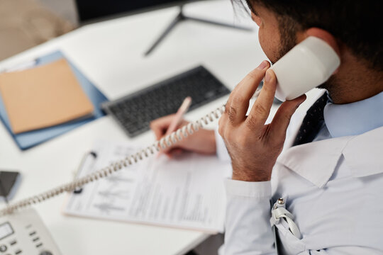 Cropped shot with focus on hand of male health practitioner holding landline phones handset and talking with patient while working at desk in clinic
