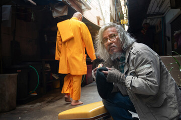 Homeless senior man sits on the sidewalk with a Chinese monk passing behind - obrazy, fototapety, plakaty