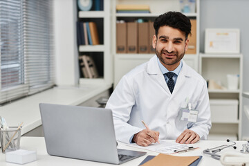 Portrait of cheerful Muslim man physician looking at camera while sitting at desk in clinic office - obrazy, fototapety, plakaty