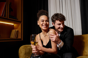 man hugging excited african american woman holding glass of wine and sitting on velvet sofa