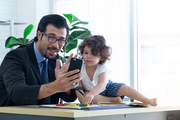 Cute little girl sitting and playing on desk at father's office, paid attention to something on the phone screen, dad uses his cell phone to make a video call with someone