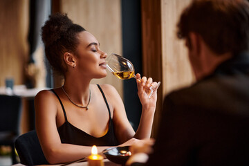 happy african american woman drinking white wine next to boyfriend during date on Valentines day