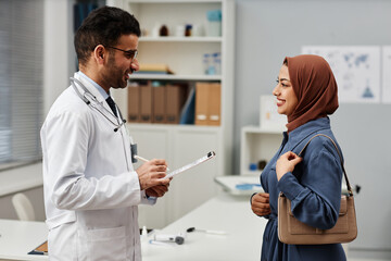 Side view at cheerful Middle Eastern male general practitioner and female patient in brown headscarf meeting in modern clinic office, doctor in glasses holding clipboard with medical card - obrazy, fototapety, plakaty