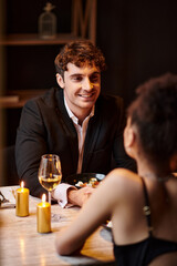 joyful man in elegant attire looking at girlfriend on blurred foreground during date in restaurant