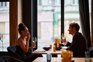 happy interracial couple in elegant attire holding glasses of wine during date in restaurant, laugh