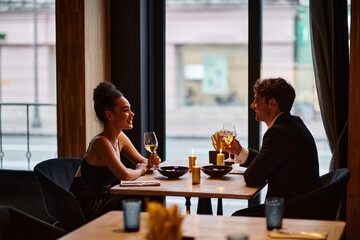 happy interracial couple in elegant attire holding glasses with wine during date in restaurant - obrazy, fototapety, plakaty