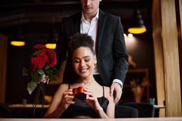 man gifting red roses to happy african american woman holding heart-shaped box on Valentine`s day
