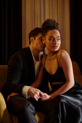 Obraz premium handsome man in suit seducing pretty african american woman in black dress and sitting on sofa