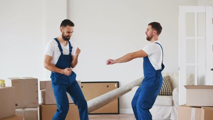 Two workers dancing at new home around cardboard boxes. Mans having fun celebrating moving in new apartment together