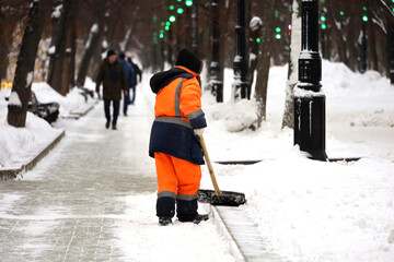 Communal services worker in uniform with a shovel clears snow on a sidewalk. Woman during snow...