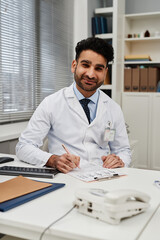 Vertical portrait of smiling Muslim male physician wearing lab coat and badge looking at camera while sitting at desk in clinic office