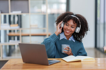 Cheerful young african american woman wearing headphones looking at laptop computer screen drinking...