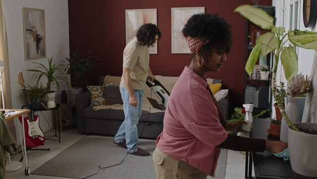 Medium Shot Of Young Interracial Couple Doing Housework Together In Modern Apartment, Biracial Woman Dusting Surfaces With Microfiber Cloth And Caucasian Man Doing Vacuuming