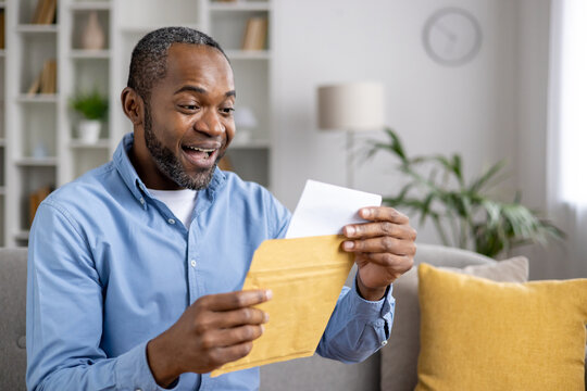 Senior joyful man sitting on sofa at home, satisfied smiling african american man reading mail message holding letter with good news in hands, inside living room of house.