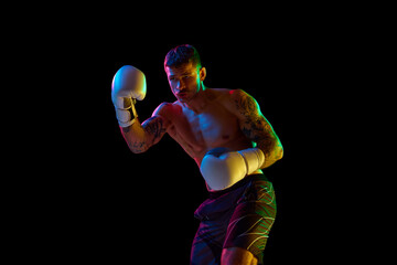 Young man with shirtless, strong, muscular body training, boxing athlete punching isolated over black background in neon light. Concept of professional sport, combat sport, martial arts, strength