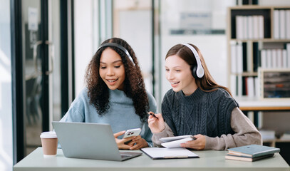 Two Attractive young female college students working on the school project using computer and tablet together, enjoy talking and headphones having a video chat.