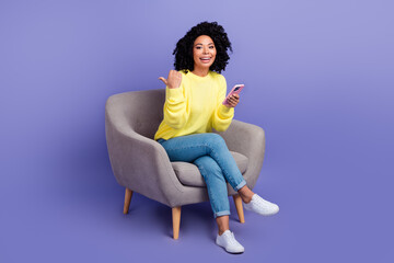 Full size photo of nice cheerful woman sit on armchair indicating at eshop empty space hold smartphone isolated on violet color background