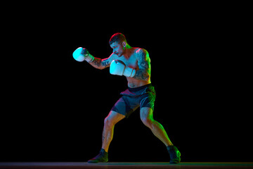 Obraz premium Full-length of shirtless young man with muscular strong body, boxer in motion, training isolated on black background in neon light. Concept of professional sport, combat sport, martial arts, strength