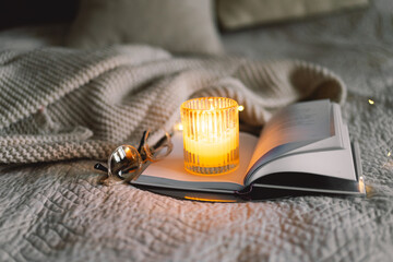 Scented candle with an open book in bed with a warm blanket. Winter holiday season. Cozy home