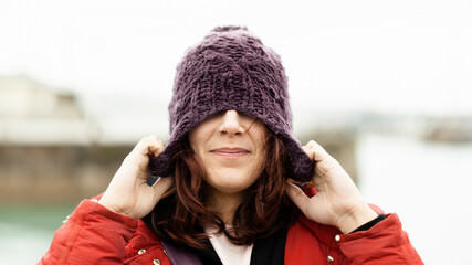 Portrait of a happy woman on the street covering her eyes with a wool hat in winter enjoying the freezing morning of the cold season. Nice woman playing with her winter hat on the street posing