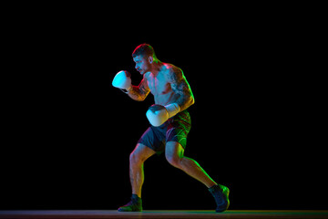 Full-length image of young athletic man, muscular, shirtless boxer in motion, fighting, practicing hooks isolated over black background in neon. Concept of sport, combat sport, martial arts, strength