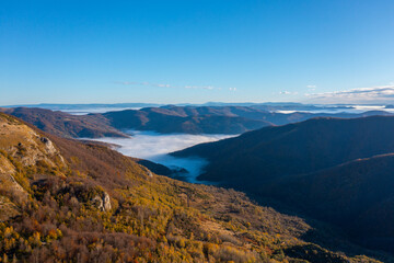 A beautiful view during autumn of a valley filled with fog and surrounded by mountain peaks and hills covered with colorful forest