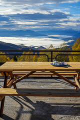 A table next to a mountain lodge with tea served and a beautiful view during autumn of a valley filled with fog and surrounded by mountain peaks and hills covered with colorful forest