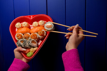 Female hands holding sushi rolls with chopsticks over a heart-shaped sushi box