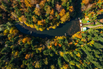 Aerial drone shot of a artificial lake with a small dam and little house near it surrounded by colorful forest during autumn.