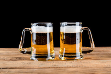 Two glass beer mugs with beer on a rough wooden table against a black background.
