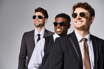 cheerful diverse friends in business casual attire with sunglasses posing together on gray backdrop