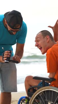 A disabled person in a wheelchair on the beach having fun with a friend and a video camera, taking a selfie