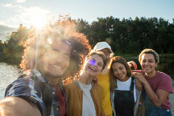 Sunshine Selfie: A A spontaneous selfie captures the exuberant spirit of a group of friends by the lakeside. The image features a Middle-Eastern man with curly hair, a young Caucasian man, an elderly