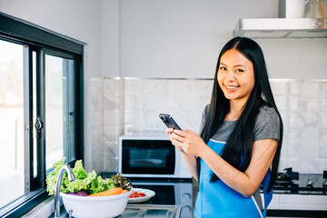 In a cozy kitchen an Asian woman cooks vegetables viewing a cooking tutorial on her smartphone....